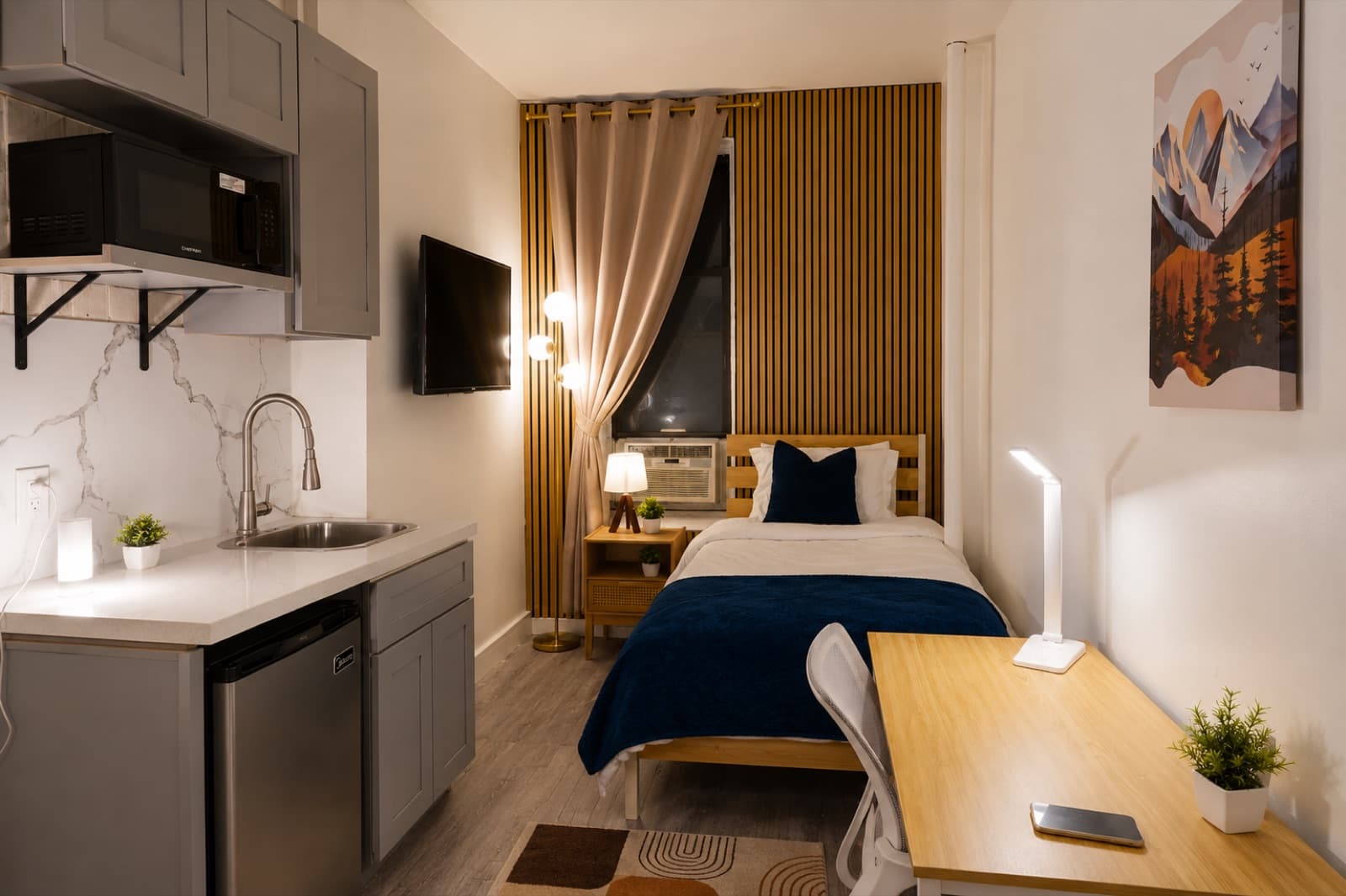 Deluxe furnished room at Amsterdam Place, Upper West Side NYC — kitchenette, bed, and wall-mounted TV
